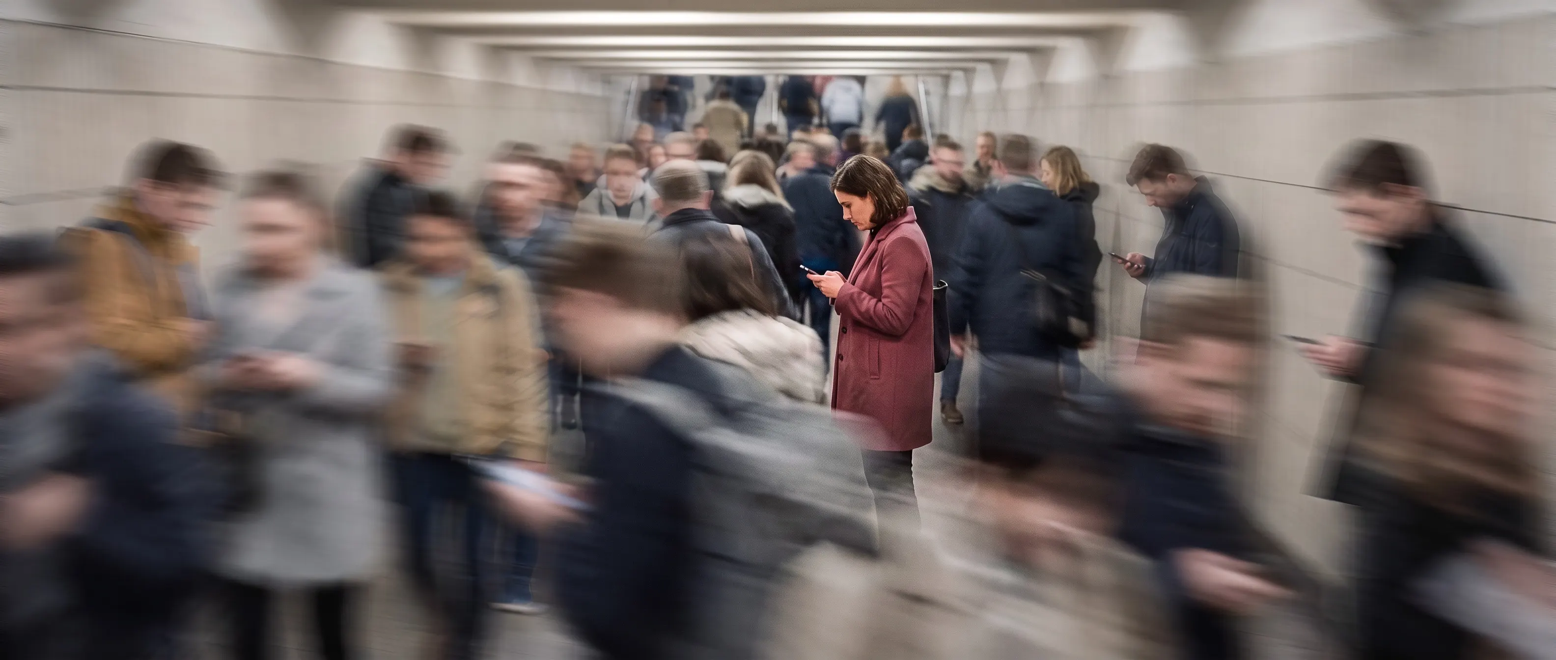 Fußgängertunnel, Pendlerströme zu Bändern verwischt — eine Frau im altrosa Mantel steht scharf am Smartphone